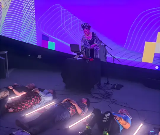 A performer wearing a sensor headset stands at a table in front of a large projection, while audience members lie on the floor among glowing LED tubes