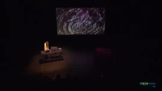 A performer seated at a table on a dark stage, under a large projected image