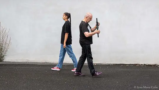 Two performers walking in opposite directions in front of a white wall, one of them holding a long digital instrument